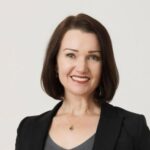 A woman with short brown hair smiles, wearing a black blazer over a gray top, against a light background.